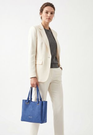 Femme en costume crème et haut gris tenant un sac à main en cuir bleu, debout devant un fond blanc uni.