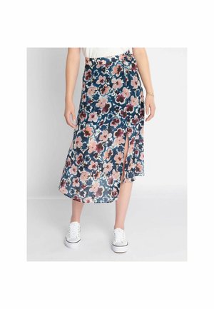 Femme portant une jupe midi fleurie avec une fente latérale, des baskets blanches et un haut blanc, debout devant un fond blanc uni.