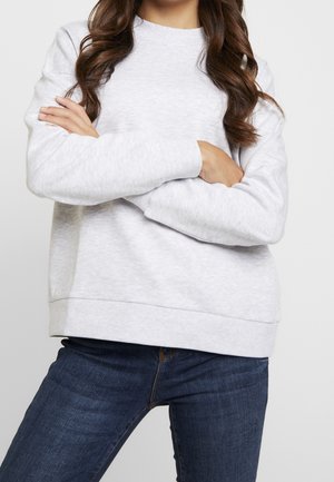 Femme portant un sweat-shirt gris clair et un jean bleu foncé, les bras croisés devant un fond uni.