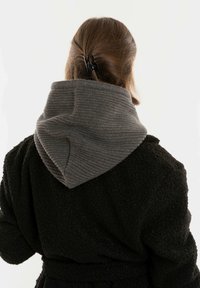 Vrouw met bruin haar dat naar achteren is gekleed, draagt een zwarte, gestructureerde jas met een grote grijze gebreide kap, gezien van achteren tegen een eenvoudige achtergrond.