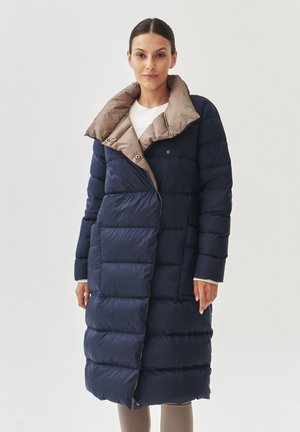 Abrigo de plumas azul marino con un cuello beige, con paneles acolchados, cierre de botones y un diseño asimétrico. Textura suave.