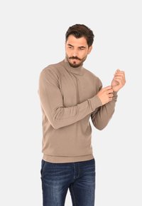Maglione beige a collo alto con maniche lunghe, polsini e orlo a coste, indossato con jeans blu. Tessuto in maglia morbido, design semplice e aderente.