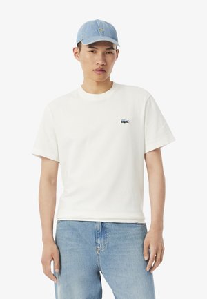 T-shirt bianca in cotone con maniche corte, con un piccolo logo Lacoste blu e verde. Abbinata a jeans di denim blu chiaro e un cappellino blu chiaro.