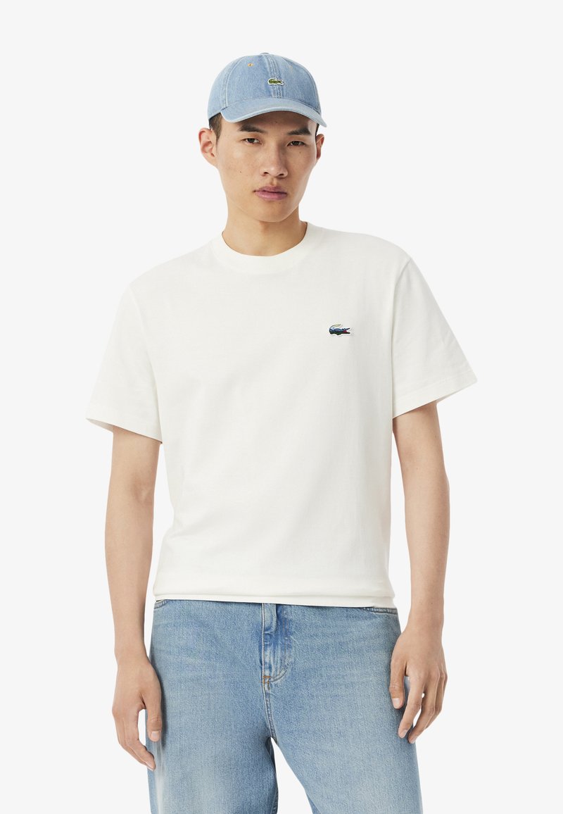T-shirt bianca in cotone con maniche corte, con un piccolo logo Lacoste blu e verde. Abbinata a jeans di denim blu chiaro e un cappellino blu chiaro.