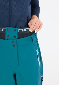 Tealfärgade skidbyxor med svart midja och knäppningar, sidofickor med dragkedja och en slät, jämn tygtextur.