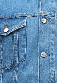 Ανοιχτό μπλε τζιν μπουφάν με ορατή ραφή, τσέπη με καπάκι και μεταλλικά κουμπιά με σήμανση που έχουν λεία υφή.