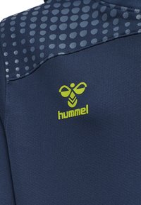 Marinefarbener Sporttop aus strukturiertem Material mit einem gepunkteten Muster auf den Schultern und einem leuchtend grünen "hummel"-Logo auf der Vorderseite.