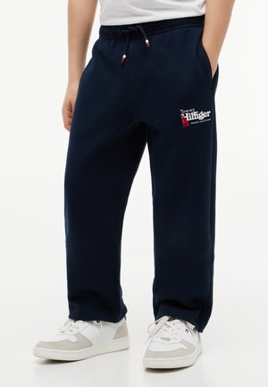 Person iført navyblå Tommy Hilfiger joggingbukser med logo på låret og hvide sneakers med grå accenter.