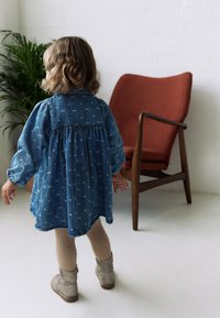 Džínové šaty so zavinutým pásom, s vzorom svetlomodrej farby. Béžové pletené pančuchy a sivé členkové čižmy. V pozadí hnedé akcentové kreslo.