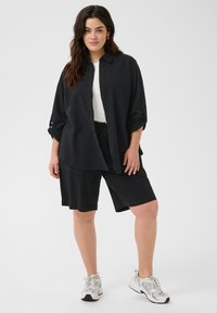 Camisa negra de botones con mangas enrolladas, combinada con shorts negros. El modelo lleva una camiseta blanca y zapatillas grises. Un conjunto casual y holgado.