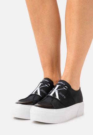 Jambes portant des baskets compensées noires sans lacets avec des brides blanches ornées du logo "CK" et des semelles épaisses blanches sur un fond blanc.
