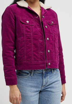 Kvinde iført en tætsiddende lilla kordfrakke med hvid sherpa-krave og sølvknapper, kombineret med lyseblå denim-jeans.