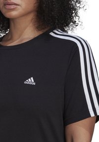 Svart bomullst-shirt med vit tre-randig design på axlarna och liten vit broderad logo på bröstet. Kort ärm, rund hals.