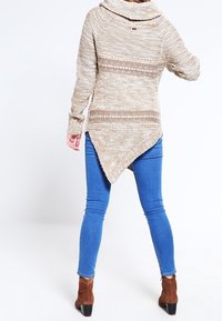Beżowy dziany sweter z golfem i asymetrycznym dołem. Zawiera fakturowane wzory i długie rękawy. Połączony z niebieskimi dopasowanymi dżinsami i brązowymi botkami.