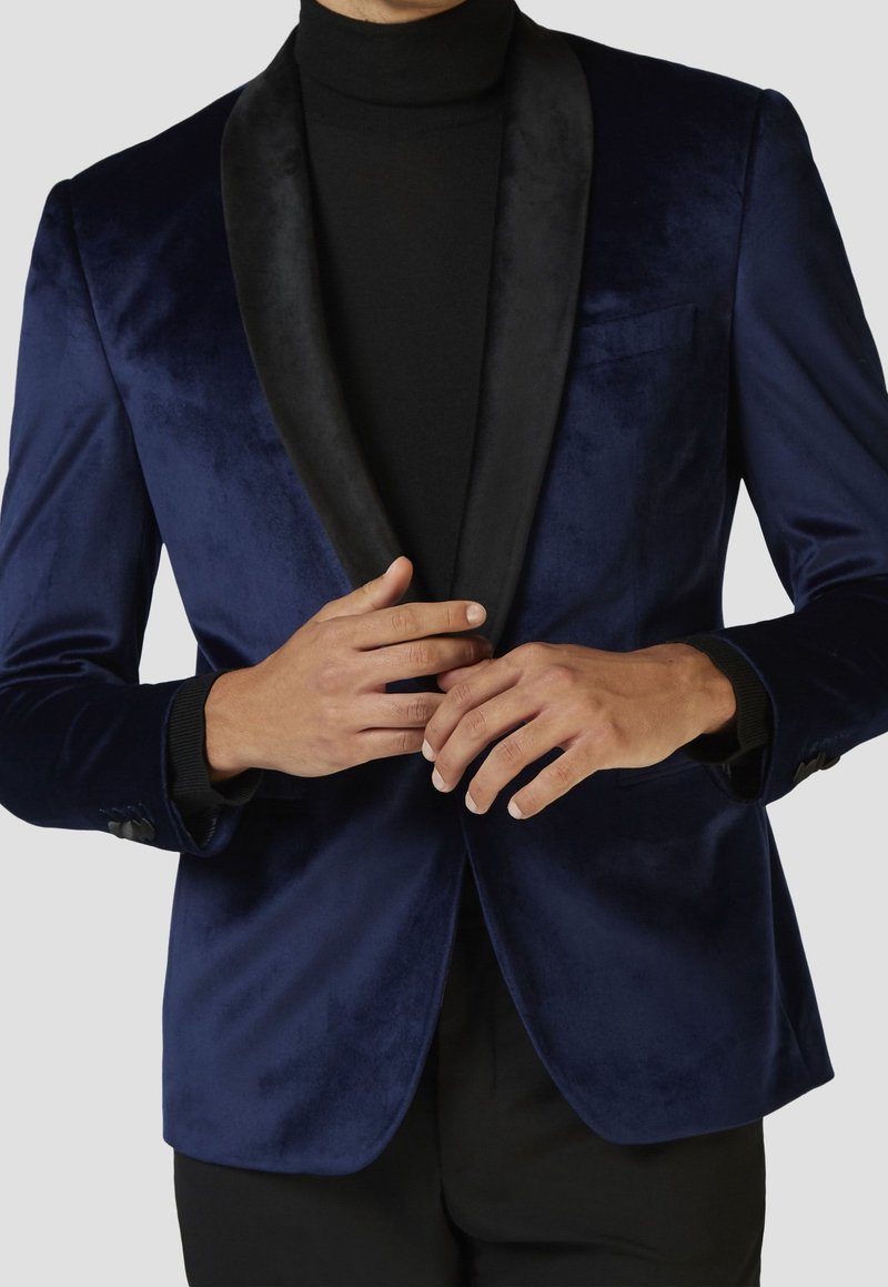Námorně modrý sametový blazers s černým saténovým šálovým límcem, jednou kapsou a knoflíkovými manžetami, nošený přes černý rolák.