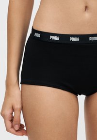 Gros plan d'une personne portant un sous-vêtement noir Puma avec une ceinture élastique de marque, montrant une partie du torse, de la main et du haut de la cuisse.