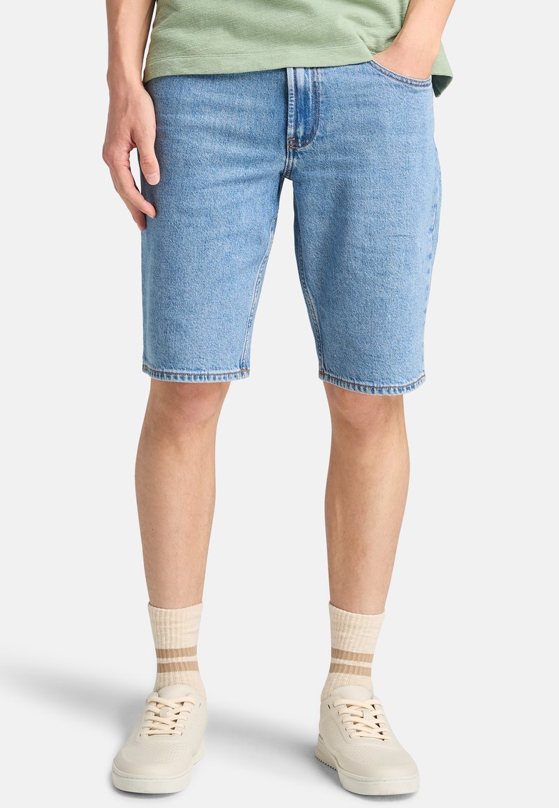 Shorts de denim azul claro con un ajuste relajado, que presentan un diseño de cinco bolsillos y costuras visibles, combinados con zapatillas crema y calcetines a rayas.