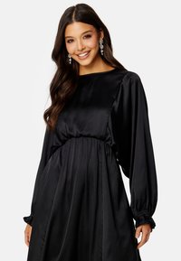 Černé saténové šaty se staženým pasem, dlouhými nabíranými rukávy a hladkou texturou. Mají kulatý výstřih a volánkové manžety.