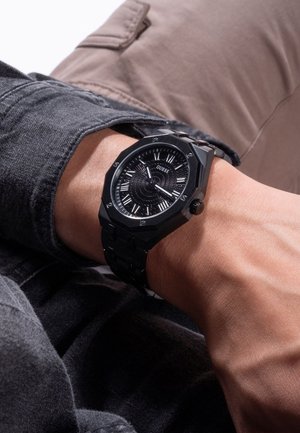 Montre octogonale noire avec un cadran noir texturé, index argentés et un bracelet en métal noir à maillons. Portée au poignet, posée sur un tissu gris.