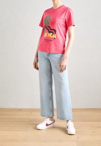 Persona vistiendo una camiseta gráfica roja con el texto "Beach", jeans de pierna ancha de color azul claro y zapatillas blancas con acentos rojos, de pie sobre un suelo de madera.