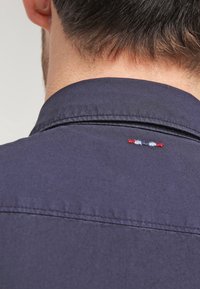 Mörkblå skjorta med nedkläppad krage, med dekorativ sömnad i rött, vitt och blått vid nackpartiet. Slät bomullstextur.