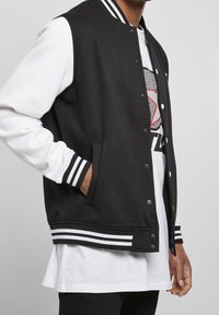 Zwarte varsityjack met witte gestreepte manchetten en zoom, met drukknoopsluiting en zijzakken. Draagt over een wit T-shirt met een grafische print.