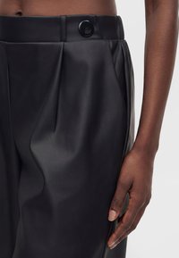 Pantalon taille haute en simili cuir noir avec un grand bouton noir, porté par une personne à la peau foncée, montrant la main droite reposant sur le côté.