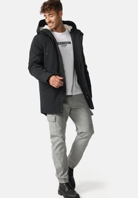 Parka noire avec une capuche en fourrure, poches boutonnées, fermetures éclair, assortie à un pantalon cargo gris et des bottes noires à lacets. Tenue décontractée.