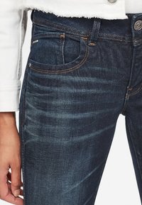 Jeans de mezclilla azul oscuro con un corte ajustado, que presentan un ligero desgaste, un diseño clásico de cinco bolsillos y costuras en contraste.