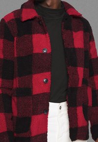 Red and black plaid fleece jacket with a collar, two front buttons, and textured fabric. Worn over a black turtleneck.