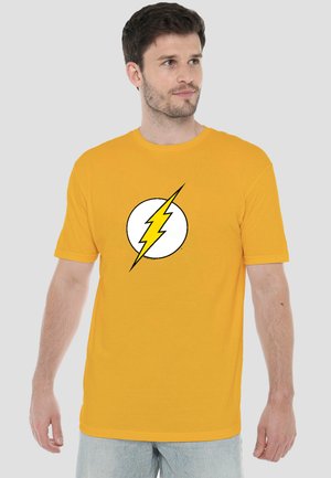 Mann trägt gelbes T-Shirt mit weißem Kreis und gelbem Blitzsymbol, schaut nach links vor einfarbigem Hintergrund.