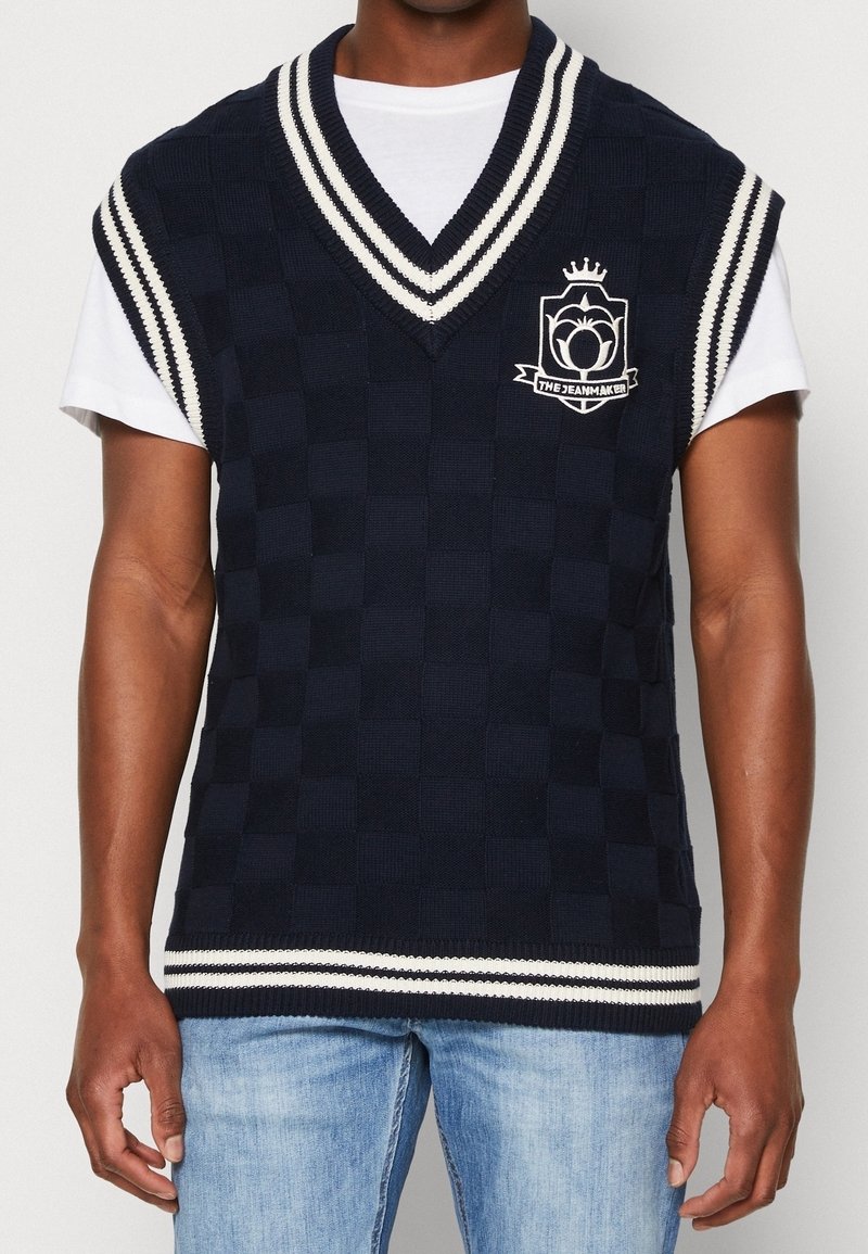 Gilet sans manches bleu marine avec un motif à carreaux, encolure en V, bordure rayée blanche et logo brodé sur la poitrine.