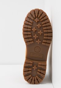 Semelle d'une botte Timberland marron avec des crampons profonds et des motifs en forme de croix, debout sur fond blanc.