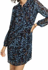 Femme portant une robe bleue à motifs, à manches longues, longueur genoux, coupe ample avec un col froncé, debout avec une main sur la hanche.