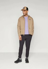 Beige Reißverschlussjacke mit Kapuze, lila T-Shirt, schwarze Sporthose und eine beige Kappe mit Logo. Schwarze Turnschuhe vervollständigen das Outfit.