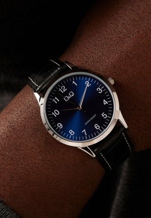 Montre-bracelet ronde avec cadran bleu, boîtier en argent, bracelet en cuir noir, chiffres blancs et aiguilles indiquant 12h10 sur un poignet à peau foncée.