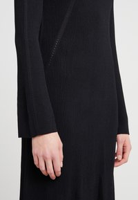 Mains et avant-bras d'une personne portant une robe noire à manches longues à côtelage avec de petits trous décoratifs sur les manches et le côté.