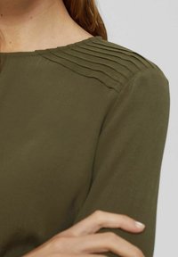 Blusa verde oliva con dettagli plissettati lungo le spalle, texture liscia e maniche lunghe.