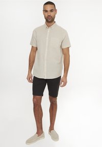 Camisetta beige a maniche corte con bottoni e taschino frontale, abbinata a pantaloni corti neri e scarpe slip-on chiare. Vestibilità slim e tessuto traspirante.