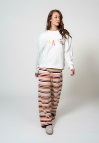 Fehér pulóver, amelyen színes "CH ÁPRIL" felirat található, széles szárú, csíkos nadrággal párosítva, rózsaszín, kék, sárga és fehér színekben. Hétköznapi lábbeli.