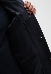 Black jacket interior with smooth satin lining, featuring a side pocket and a visible stitching detail.  Hand gripping the edge of the jacket.