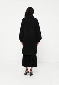 Cappotto oversize nero con superficie testurizzata, maniche ampie e colletto, indossato sopra una gonna nera plissettata e sandali con tacco.