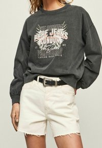 Sudadera gris oscuro con texto gráfico en blanco y rosa, cuello redondo y mangas largas. Combinada con pantalones cortos de mezclilla beige claro con dobladillo desflecado.
