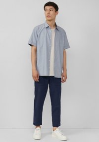 Kurzärmliges, hellblaues, knopfbares Hemd, getragen über einem cremefarbenen T-Shirt, kombiniert mit navyblauen Hosen und weißen Turnschuhen. Einfaches Design mit minimalen Details.