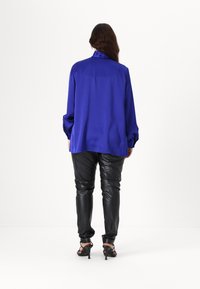 Satin blå bluse med høj krave og lange, voluminøse ærmer, parret med tætsiddende sorte læderbukser og stropede hæle.