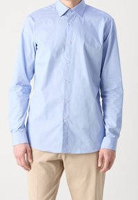 Camicia azzurra chiaro a maniche lunghe con una texture liscia, colletto a punta e bottoni bianchi, abbinata a pantaloni beige.