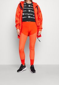 Jaskrawo-pomarańczowy zestaw odzieży sportowej z krótki topem z czarnym wzorem logo, pasującymi legginsami z teksturowanymi panelami oraz czarnymi butami sportowymi.