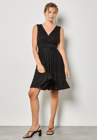 Czarna sukienka bez rękawów z dekoltem w serek, zapinana na krzyż, z teksturowanej tkaniny i wiązaniem w talii. Połączona z czarnymi sandałami na szpilkach.