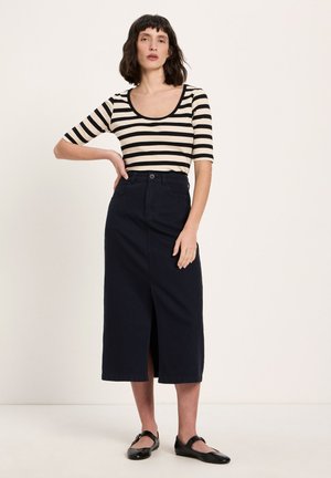 Frau steht mit einer Hand in der Hüfte, trägt ein schwarz-weiß gestreiftes Oberteil, einen schwarzen Midirock mit vorderlichem Schlitz und schwarze Ballerinas.