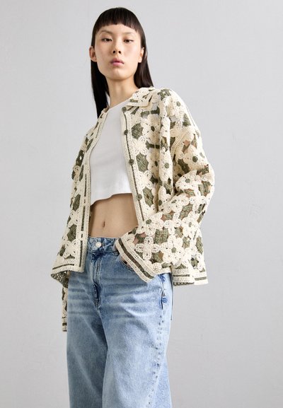 Heklet jakke i kremfarge med grønt blomsterdesign, åpen foran, med ribbestrikket kant. Bært over en hvit cropped tank top og lyseblå jeans.